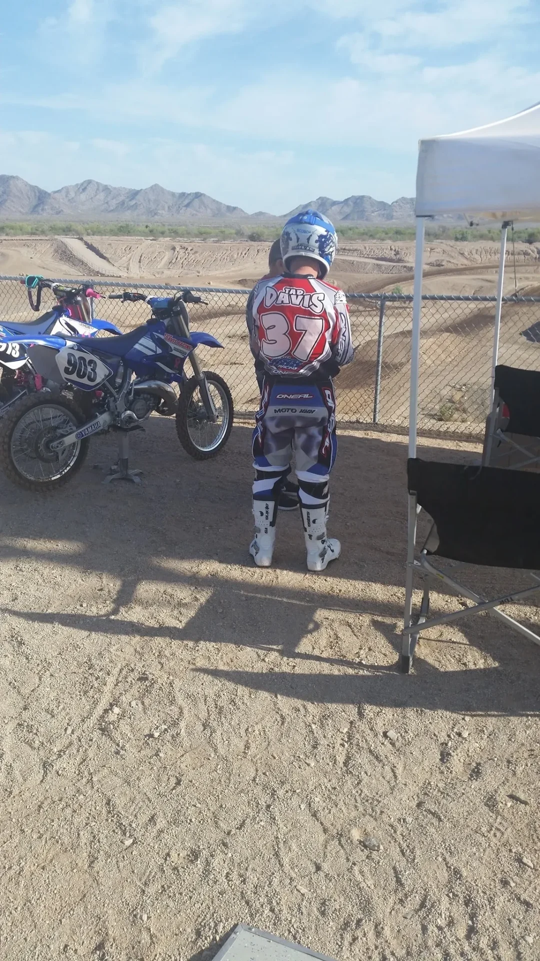 A person standing in dirt with a motorcycle behind him.