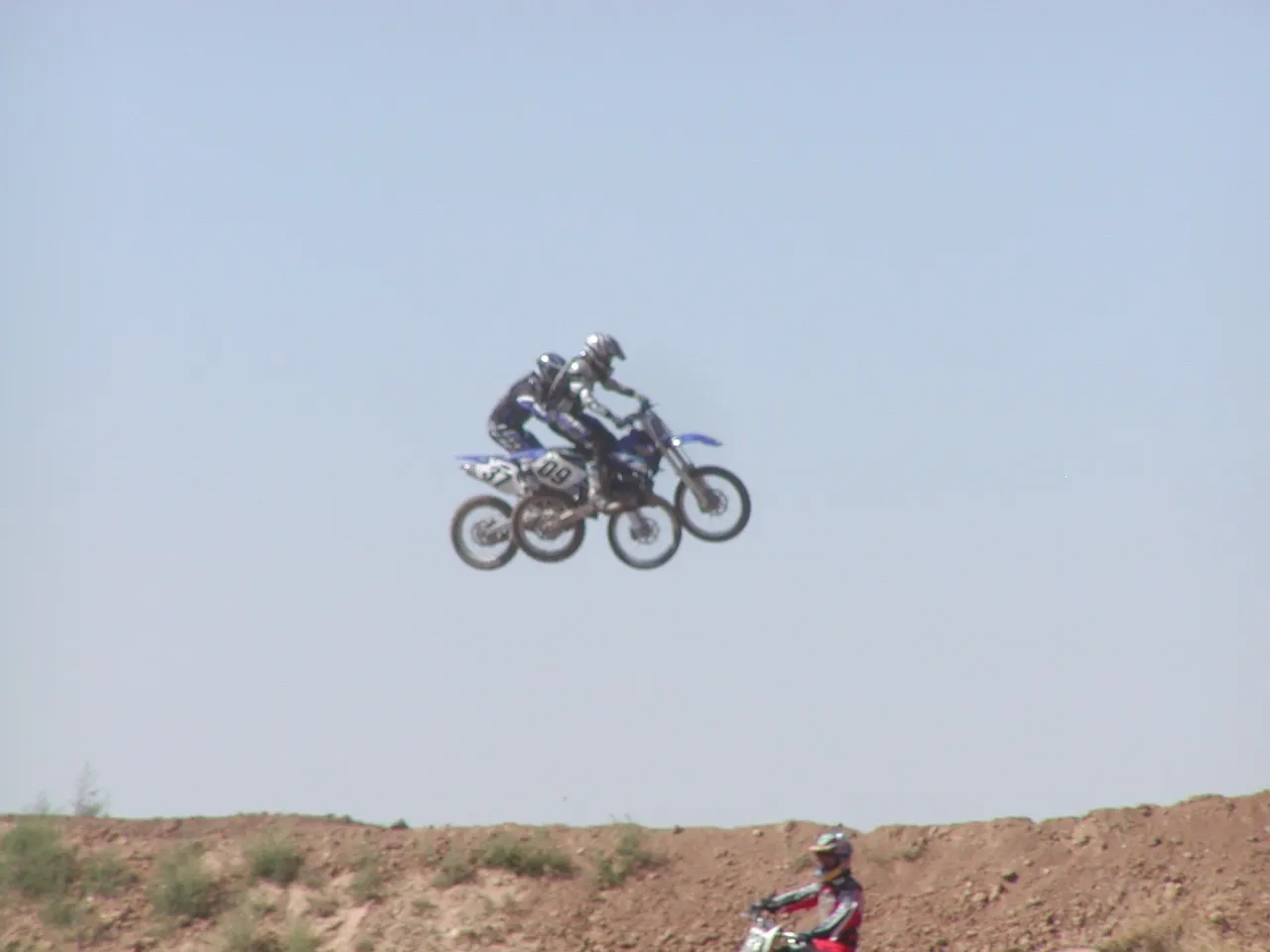 Two people on motorcycles are jumping over a hill.